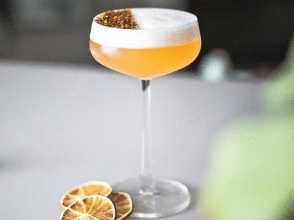 Elegant cocktail glass with a creamy Elderberry Sour, garnished with herbs on the foam and dried lime slices on the table.