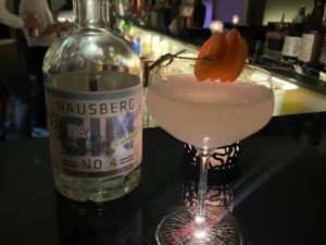 Hausberg No.4 French 75 cocktail served in an elegant coupe glass, garnished with orange slices, next to a bottle of Hausberg Gin in a dimly lit bar setting.