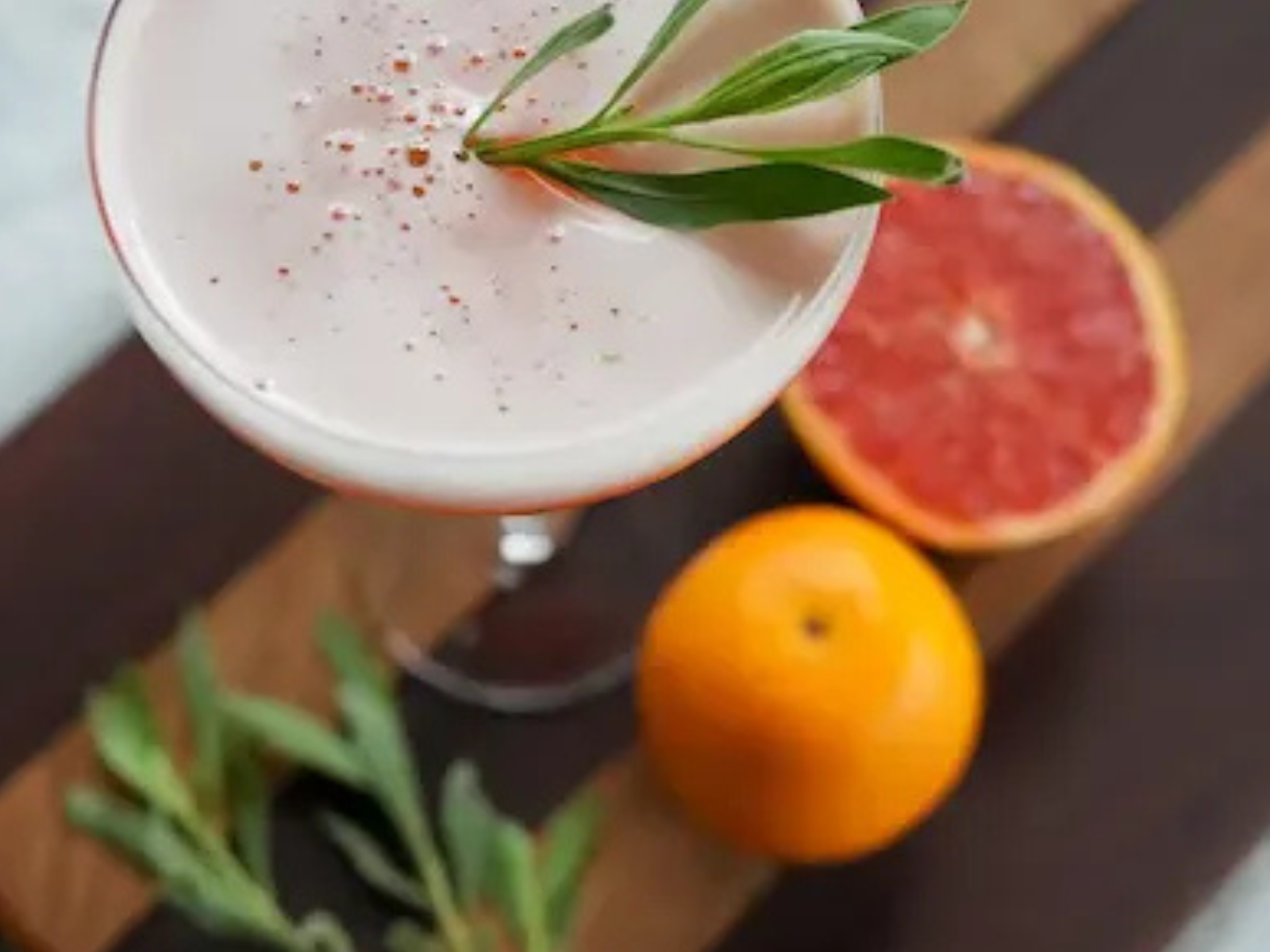 Okavango Sunset , an elegant gin cocktail in a coupe glass, garnished with wild sage, with grapefruit and orange in the background.