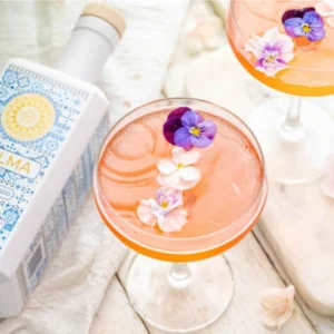 Palma Cosmopolitan cocktail served in a coupe glass with edible flower garnish, next to a bottle of Palma gin.