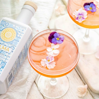 Palma Cosmopolitan cocktail served in a coupe glass with edible flower garnish, next to a bottle of Palma gin.